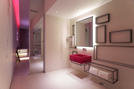 A modern bathroom with a sleek and minimalist design. It features a bold red sink mounted onto the wall below a large mirror that has a backlight. The wall next to the sink is decorated with various rectangular metal frame pieces of different sizes. There is a shelf below the sink holding rolled white towels. The room has a subtle and elegant lighting with a notable red accent light on the ceiling extending through the hallway. The floor is covered in light-colored tiles and the overall space has a contemporary and artistic feel with smooth surfaces and clean lines.