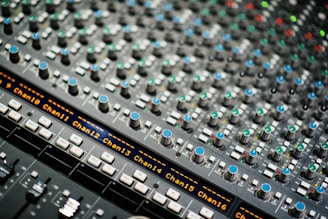 A close-up view of a complex audio mixing console with multiple knobs, buttons, and sliders. The console features blue-topped knobs, small lights, and digital displays showing channel numbers.