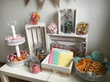 A festive candy and snack display is arranged on a table. Various jars filled with colorful candies, including gummies, chocolates, and marshmallows, are placed on wooden crates. A two-tiered tray holds wrapped candies, while a bowl contains chips and savory snacks. Decorative napkins in polka dot and striped patterns add a playful touch to the setup.