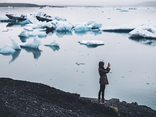 woman taking photo standing near body of water in Jökulsárlón Iceland