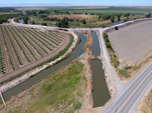 Aerial view of a rural landscape featuring neatly organized agricultural fields and a network of irrigation canals. The scene includes a narrow paved road intersecting the fields and waterways, with a bridge crossing over one of the canals. The fields are partially cultivated, and the landscape is surrounded by greenery and distant hills.