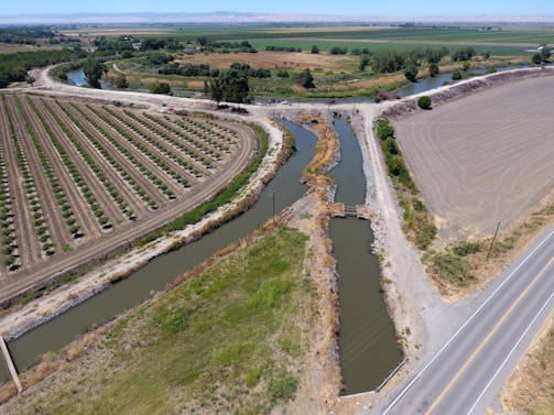 Aerial view of a rural landscape featuring neatly organized agricultural fields and a network of irrigation canals. The scene includes a narrow paved road intersecting the fields and waterways, with a bridge crossing over one of the canals. The fields are partially cultivated, and the landscape is surrounded by greenery and distant hills.