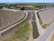 Aerial view of a rural landscape featuring neatly organized agricultural fields and a network of irrigation canals. The scene includes a narrow paved road intersecting the fields and waterways, with a bridge crossing over one of the canals. The fields are partially cultivated, and the landscape is surrounded by greenery and distant hills.