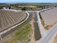 Aerial view of a rural landscape featuring neatly organized agricultural fields and a network of irrigation canals. The scene includes a narrow paved road intersecting the fields and waterways, with a bridge crossing over one of the canals. The fields are partially cultivated, and the landscape is surrounded by greenery and distant hills.