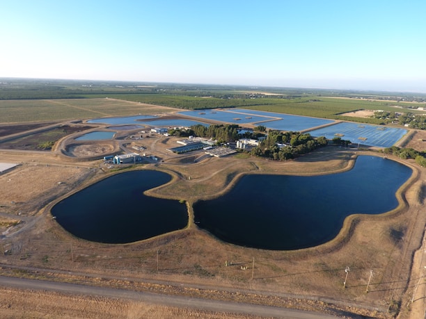 An aerial view showcasing a landscape with large water bodies, possibly reservoirs or ponds, surrounded by agricultural fields. There are solar panels installed in rows, several long buildings possibly used for processing, and clusters of trees providing greenery amid predominantly dry terrain.