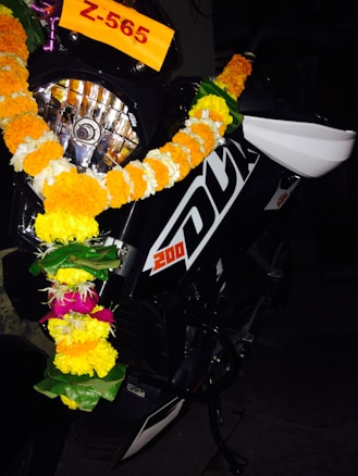 A motorcycle decorated with a garland made of bright yellow, white, and pink flowers. The motorcycle has the number '200' and 'DUK' written on its black body. A number plate with the alphanumeric code 'Z-565' is visible near the handlebar.