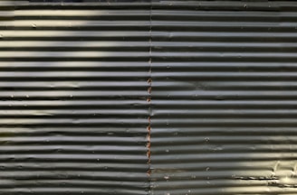 Corrugated metal sheets are aligned vertically with visible screws and a central seam. The surface appears weathered with some discoloration and shadows cast across it.