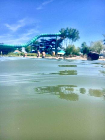 A green water slide with a twisting structure is visible in the background. The photo is taken from a low angle close to the water surface of a pool or lake. There are people standing near the water's edge, and trees can be seen surrounding the area under a clear blue sky.