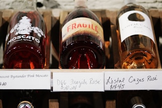 Three bottles of rosé wine are displayed horizontally on a wooden shelf. The labels on the bottles are prominently visible, with one featuring the word 'FRAGILE' in large red letters.
