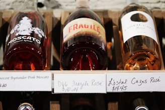 Three bottles of rosé wine are displayed horizontally on a wooden shelf. The labels on the bottles are prominently visible, with one featuring the word 'FRAGILE' in large red letters.