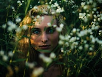 A close-up portrait of a person with prominent blue eyes and light blonde hair, partially obscured by tall green grass and small white flowers. The sunlight casts soft shadows across the face, creating an intimate and slightly mysterious atmosphere.