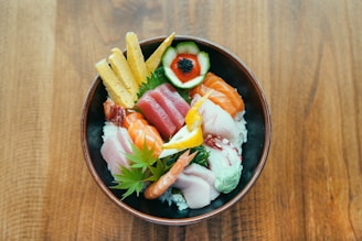 A beautifully arranged bowl of sushi ingredients including salmon, tuna, shrimp, cucumber, egg, and a leaf garnish atop a bed of rice. The vibrant colors of the fresh ingredients contrast with the dark bowl and wooden table beneath.