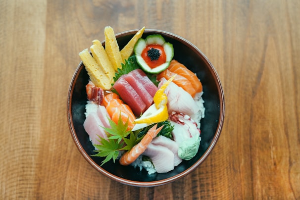 A beautifully arranged bowl of sushi ingredients including salmon, tuna, shrimp, cucumber, egg, and a leaf garnish atop a bed of rice. The vibrant colors of the fresh ingredients contrast with the dark bowl and wooden table beneath.