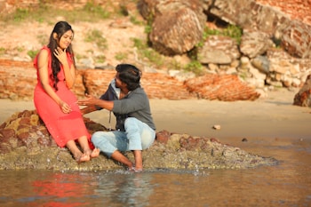A man and a woman are sitting on a rocky outcrop by the water. The woman is wearing a bright coral dress and appears contemplative, while the man, dressed in a casual shirt and jeans, is engaged in conversation, gesturing expressively. The background features an old brick wall and a sandy beach.
