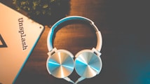 A pair of silver and white over-ear headphones rests on a dark wooden surface. Near the headphones, there is a corner of a metallic object, possibly a tablet or laptop, with the word 'Unsplash' visible on it. The setting appears to be well-lit but has a warm and relaxed ambiance with minimal elements, including some greenery in the upper left corner.