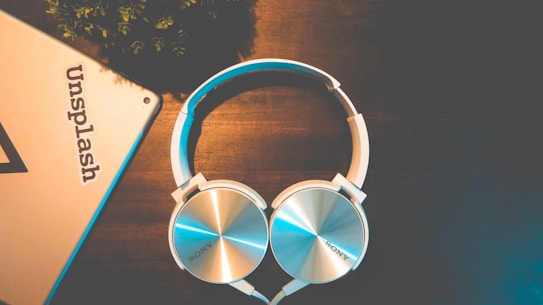A pair of silver and white over-ear headphones rests on a dark wooden surface. Near the headphones, there is a corner of a metallic object, possibly a tablet or laptop, with the word 'Unsplash' visible on it. The setting appears to be well-lit but has a warm and relaxed ambiance with minimal elements, including some greenery in the upper left corner.