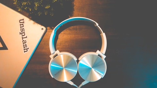 A pair of silver and white over-ear headphones rests on a dark wooden surface. Near the headphones, there is a corner of a metallic object, possibly a tablet or laptop, with the word 'Unsplash' visible on it. The setting appears to be well-lit but has a warm and relaxed ambiance with minimal elements, including some greenery in the upper left corner.