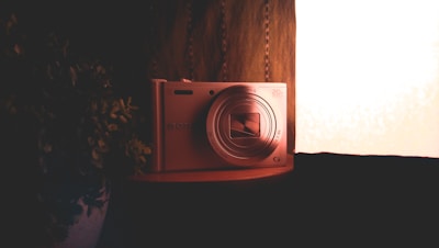 A digital camera with a prominent lens is placed against a warm background. The scene is dimly lit with soft lighting highlighting the camera's metallic surface. Beside the camera, a plant with dark leaves adds contrast.