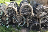 A stack of black and weathered fishing traps, possibly lobster pots, arranged outdoors on grassy terrain. The traps are constructed with durable netting and have various ropes and plastic components attached.