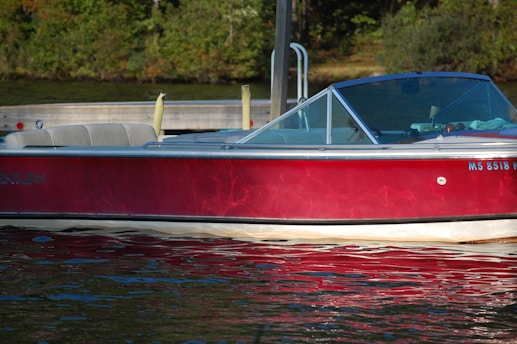 A red motorboat is docked near a wooden pier surrounded by calm water. The glistening water reflects light onto the side of the boat, giving it a shimmering appearance. The seats inside the boat are grey and well-cushioned. Dense greenery is visible in the background.