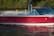 A red motorboat is docked near a wooden pier surrounded by calm water. The glistening water reflects light onto the side of the boat, giving it a shimmering appearance. The seats inside the boat are grey and well-cushioned. Dense greenery is visible in the background.