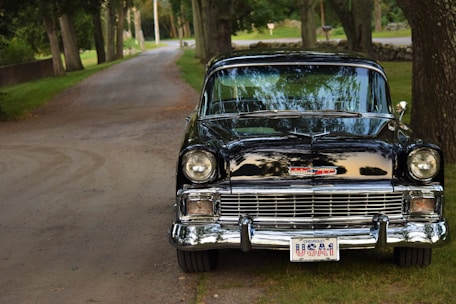A classic black Chevrolet with a polished chrome finish is parked on a dirt path lined with tall trees. The vintage car has a prominent front grille and distinctive headlights. Its license plate reads 'USA1', and reflections of the surrounding landscape are visible on the car's shiny surface. The scene is serene with green grass and trees creating a peaceful backdrop.