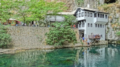 A serene view of a white building with black-framed windows situated by the edge of a clear, reflective body of water. The building is surrounded by lush green trees and set against a rocky cliff. People are seen near the building, some on a balcony overlooking the water, suggesting a relaxed, communal atmosphere.