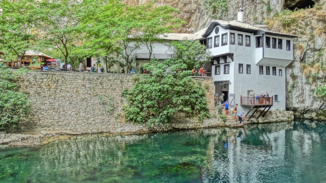 A serene view of a white building with black-framed windows situated by the edge of a clear, reflective body of water. The building is surrounded by lush green trees and set against a rocky cliff. People are seen near the building, some on a balcony overlooking the water, suggesting a relaxed, communal atmosphere.