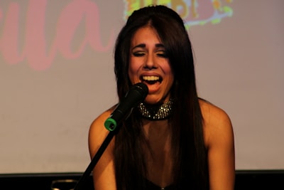A woman with long dark hair is singing passionately into a microphone. She is wearing a sparkly choker and appears to be on stage. The background is dimly lit, possibly with a blurred projection of colorful text.