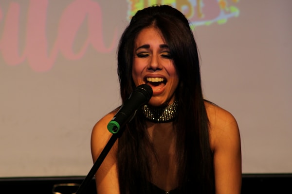 A woman with long dark hair is singing passionately into a microphone. She is wearing a sparkly choker and appears to be on stage. The background is dimly lit, possibly with a blurred projection of colorful text.