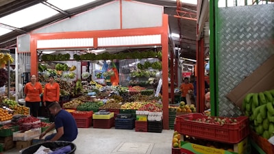 A bustling fruit and vegetable market stall displaying an abundant variety of produce. Colorful fruits and vegetables are neatly arranged in crates, with bananas hanging prominently overhead. Three individuals in orange uniforms are visible, interacting with the produce and customers. The setting is under a covered space with a metal roof and bright lighting.
