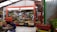 A bustling fruit and vegetable market stall displaying an abundant variety of produce. Colorful fruits and vegetables are neatly arranged in crates, with bananas hanging prominently overhead. Three individuals in orange uniforms are visible, interacting with the produce and customers. The setting is under a covered space with a metal roof and bright lighting.