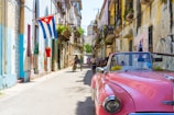 A vibrant street scene with an old pink convertible car parked on a narrow road lined with colorful, deteriorating buildings. A Cuban flag hangs from one of the buildings, and several people are seen walking or riding bicycles. The atmosphere is lively, with the architecture reflecting a rich, cultural history.