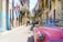A vibrant street scene with an old pink convertible car parked on a narrow road lined with colorful, deteriorating buildings. A Cuban flag hangs from one of the buildings, and several people are seen walking or riding bicycles. The atmosphere is lively, with the architecture reflecting a rich, cultural history.