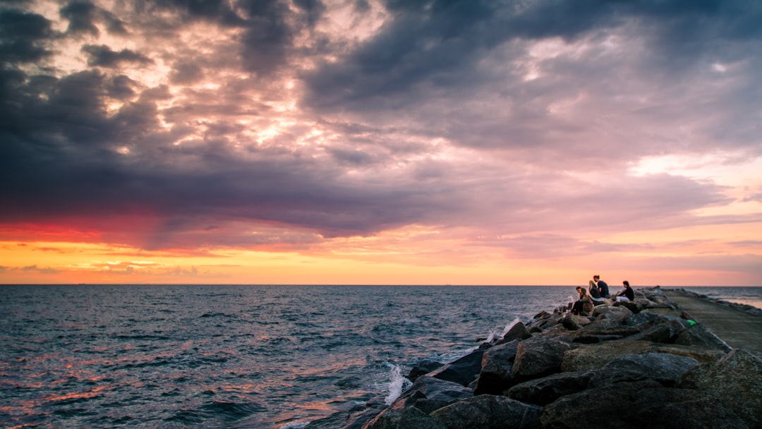 people sitting on rock near body of water, Coloured sunset at sea with people sitting on pier