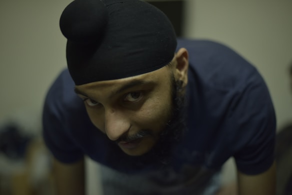 A person is leaning forward and looking directly at the camera. They are wearing a dark turban and have a beard. The background is blurred, suggesting a focus on the subject.