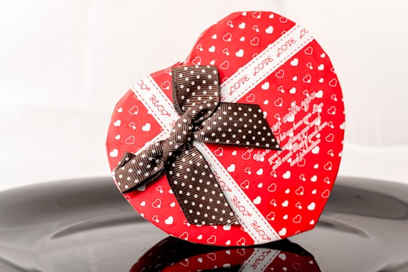 A heart-shaped gift box is prominently displayed, featuring a vibrant red color with small white heart patterns. The box is adorned with a brown ribbon with white polka dots, tied into a decorative bow. The words 'Love' and a message are printed in white on the box. The surface on which the box rests has a reflective quality, casting a slight reflection.