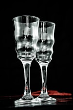 Two elegant, clear glass goblets with a textured, wavy surface are positioned side by side against a dark background. Subtle reflections and highlights add depth to the transparent material, creating a sophisticated and refined atmosphere.