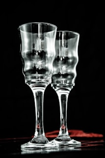 Two elegant, clear glass goblets with a textured, wavy surface are positioned side by side against a dark background. Subtle reflections and highlights add depth to the transparent material, creating a sophisticated and refined atmosphere.