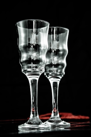 Two elegant, clear glass goblets with a textured, wavy surface are positioned side by side against a dark background. Subtle reflections and highlights add depth to the transparent material, creating a sophisticated and refined atmosphere.