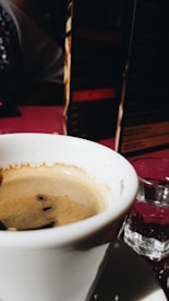 A close-up of a cup filled with coffee, placed on a saucer. The coffee appears to have a creamy texture on top. In the background, a menu with text is slightly out of focus, creating a bokeh effect. A glass of water is positioned next to the coffee cup, catching some of the ambient light.