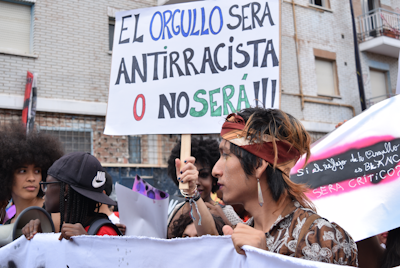 A group of people gathered in a street holding signs with messages advocating against racism. The main sign in the image is in Spanish and highlights the importance of anti-racism. The setting appears to be urban, with brick buildings visible in the background.