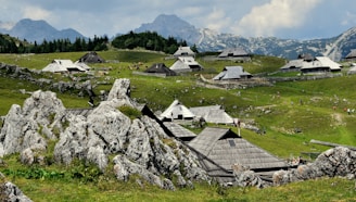 A picturesque landscape featuring traditional wooden huts scattered across a lush green hillside, with rugged rocks in the foreground. In the background, dense forests and towering mountains provide a stunning backdrop under a partly cloudy sky. Small groups of people can be seen walking around, adding life to the serene setting.