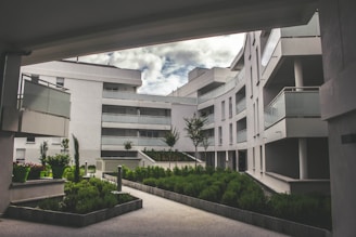 A modern residential complex featuring curved architecture with multiple stories. The buildings are white, with glass balustrades on the balconies. The setting includes landscaped green areas with well-maintained shrubs and small trees. The sky is partially cloudy, creating a serene and tranquil environment.