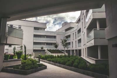 A modern residential complex featuring curved architecture with multiple stories. The buildings are white, with glass balustrades on the balconies. The setting includes landscaped green areas with well-maintained shrubs and small trees. The sky is partially cloudy, creating a serene and tranquil environment.