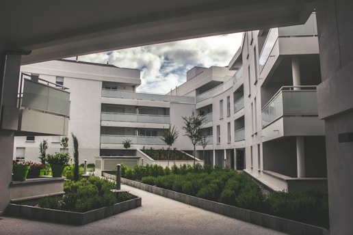 A modern residential complex featuring curved architecture with multiple stories. The buildings are white, with glass balustrades on the balconies. The setting includes landscaped green areas with well-maintained shrubs and small trees. The sky is partially cloudy, creating a serene and tranquil environment.