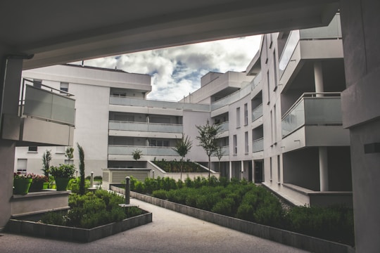 A modern residential complex featuring curved architecture with multiple stories. The buildings are white, with glass balustrades on the balconies. The setting includes landscaped green areas with well-maintained shrubs and small trees. The sky is partially cloudy, creating a serene and tranquil environment.