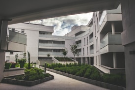 A modern residential complex featuring curved architecture with multiple stories. The buildings are white, with glass balustrades on the balconies. The setting includes landscaped green areas with well-maintained shrubs and small trees. The sky is partially cloudy, creating a serene and tranquil environment.