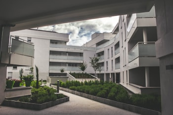 A modern residential complex featuring curved architecture with multiple stories. The buildings are white, with glass balustrades on the balconies. The setting includes landscaped green areas with well-maintained shrubs and small trees. The sky is partially cloudy, creating a serene and tranquil environment.