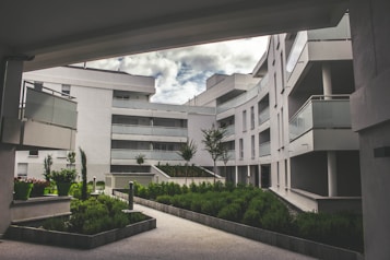 A modern residential complex featuring curved architecture with multiple stories. The buildings are white, with glass balustrades on the balconies. The setting includes landscaped green areas with well-maintained shrubs and small trees. The sky is partially cloudy, creating a serene and tranquil environment.